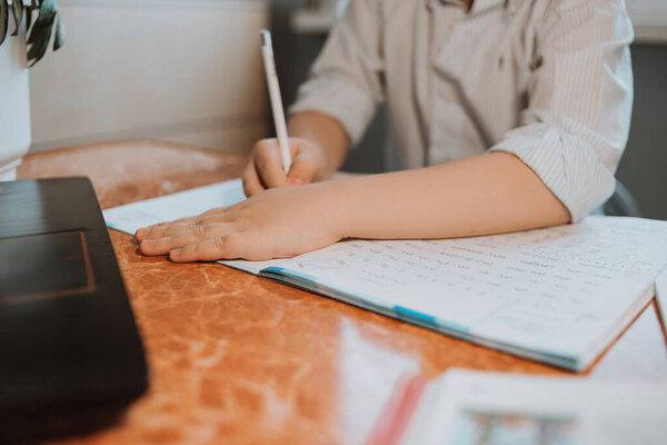 the boy writes homework in a notebook at home during CoVid-19 quarantine, distance learning online with a laptop, a child doing homework for school. Children stayed at home. Boy writes in Cyrillic.