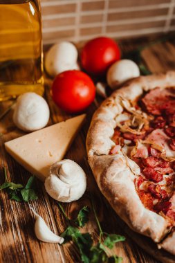 Arka planda sosisli pizza, domates, sarımsak, mantar ve şeffaf bir şişe zeytinyağı.