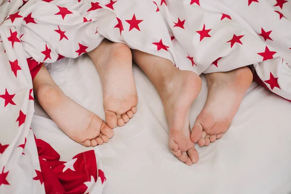 Kids\'s feet under white blanket with stars side view. Beautiful children\'s feet on the bed. Woman feet under white blanket side view. Beautiful young woman feet with blue pedicure on the bed.