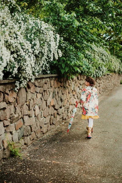 Beautiful girl in a bright raincoat walks after the rain in the park. Excellent spirea flowering bushes. Soft focus. Happy childhood concept. Young child enjoying a rainy day.
