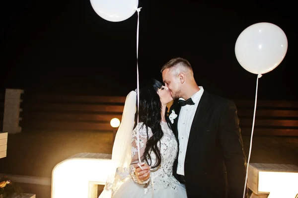 Stylish wedding couple with lights balloons stay and kisses near