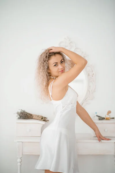 Lockiges blondes Mädchen in einem weißen Seidenkleid Boudoir-Robe auf dem Bett. — Stockfoto