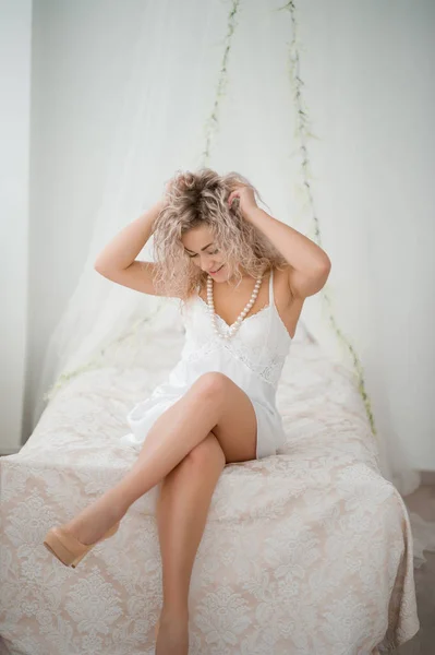 Ragazza bionda riccia in abito bianco di seta vestaglia boudoir sul letto . — Foto Stock