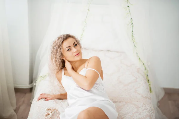 Σγουρό ξανθό κορίτσι σε μια λευκή εσθήτα μετάξι ρόμπα μπουντουάρ στο κρεβάτι. — Φωτογραφία Αρχείου