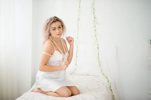 Fille blonde bouclée dans une robe de soie blanche robe de boudoir sur le lit . — Photo