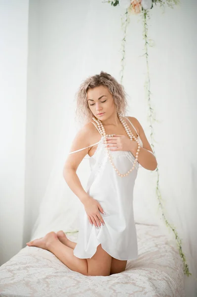 Ragazza bionda riccia in abito bianco di seta vestaglia boudoir sul letto . — Foto Stock