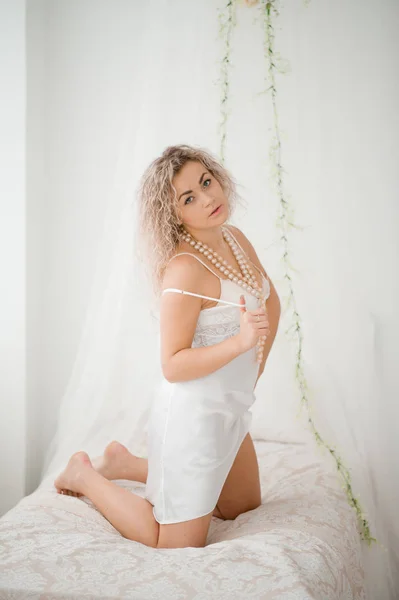 Curly blonde girl in a white silk gown boudoir robe on bed. — Stock Photo, Image
