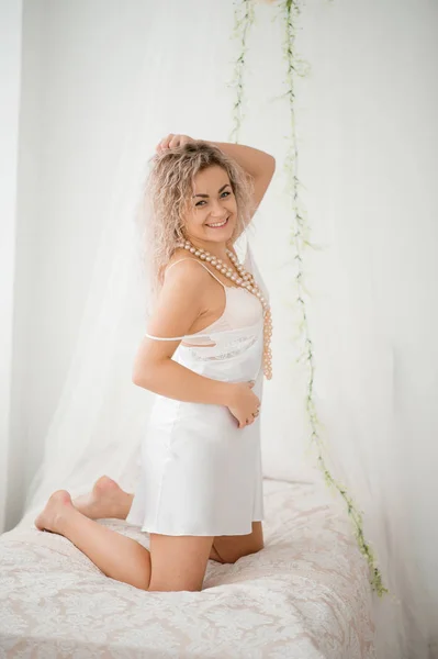ベッドの上の白い絹のドレス私室ローブで巻き毛のブロンドの女の子. — ストック写真