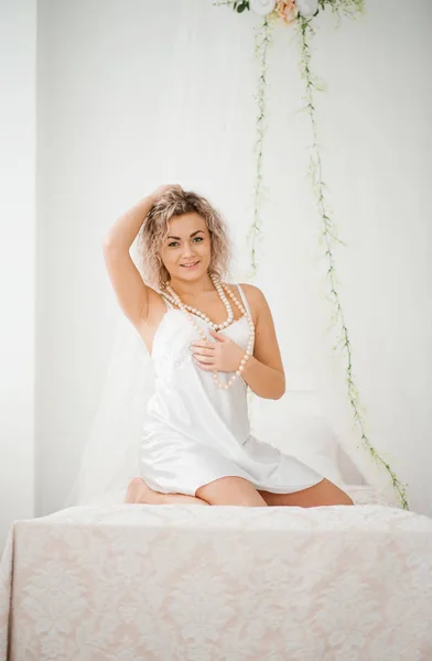 Kudrnaté blond dívka v roucho bílé hedvábné šaty budoár na posteli. — Stock fotografie