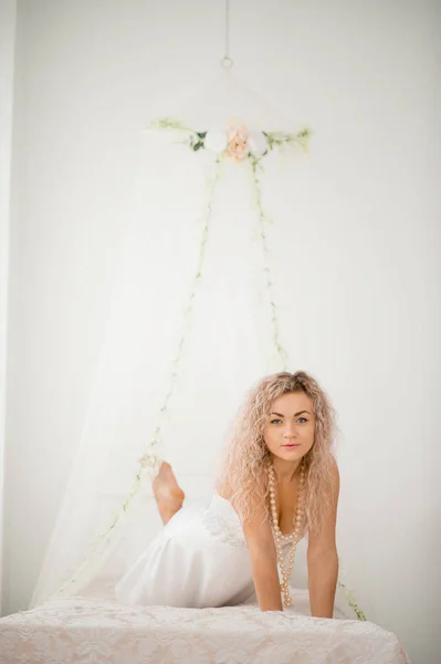 Fille blonde bouclée dans une robe de soie blanche robe de boudoir sur le lit . — Photo