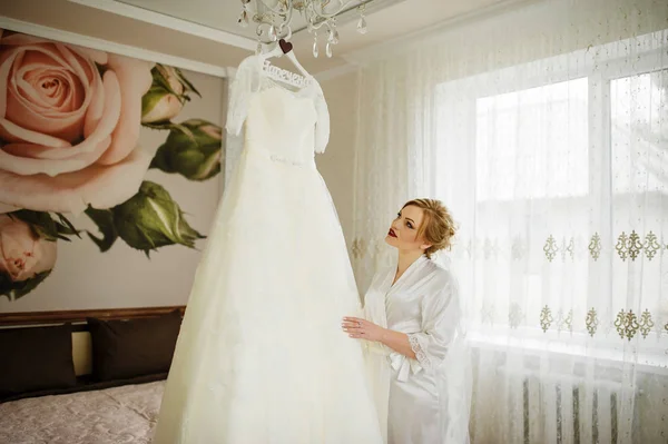 バスローブ姿で自分の部屋でウェディング ドレスを見て金髪の花嫁. — ストック写真