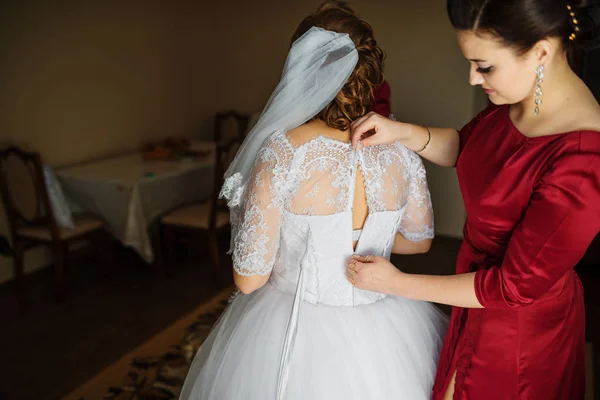 Покоївка нареченої в червоних сукнях допоможе нареченій одягнутися в весільну сукню . — стокове фото
