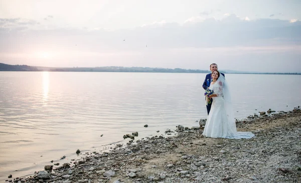 Recién casados enamorados cerca del río en la puesta del sol en su boda impresionante —  Fotos de Stock