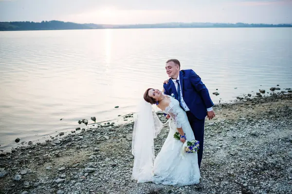 Recién casados enamorados cerca del río en la puesta del sol en su boda impresionante —  Fotos de Stock