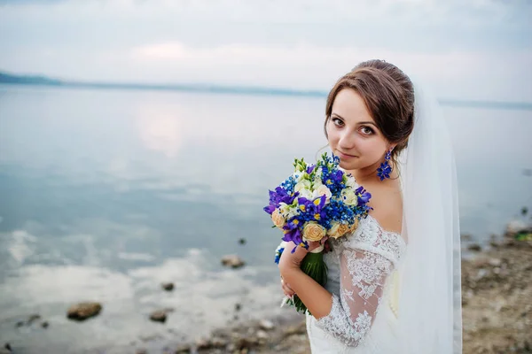 Brunette bride with violet wedding bouquet near beach of river a — Stock Photo, Image