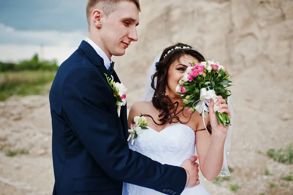 Jonge stijlvolle bruidspaar tegen zandstrand carrière bij bewolkte hemel. — Stockfoto