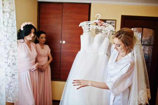 Noiva em roupão de seda olhando em seu vestido de noiva contra bridesma — Fotografia de Stock