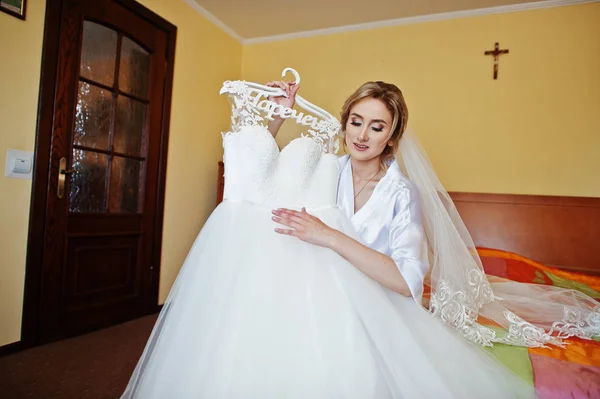 Наречена на шовковому платті дивиться на її весільну сукню, що сидить на ліжку — стокове фото
