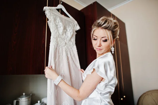 Blonde bride on silk robe with wedding dress on hangers. Morning