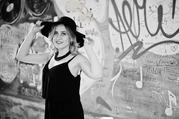 Blond kvinna på svart klänning, halsband och hat mot graffiti — Stockfoto