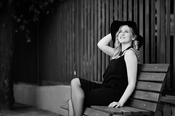 Blond kvinna på svart klänning, halsband och hatt som sitter på bänken. — Stockfoto