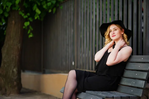 Mulher loira em vestido preto, colares e chapéu sentado no banco . — Fotografia de Stock