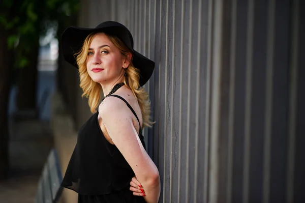 Mujer rubia en vestido negro, collares y sombrero contra cerca de hierro — Foto de Stock