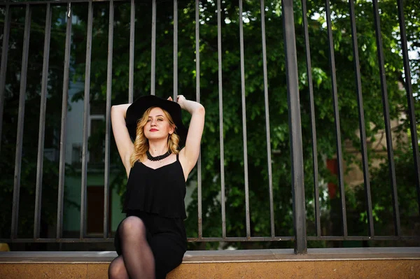 Mujer rubia en vestido negro, collares y sombrero contra cerca de hierro — Foto de Stock