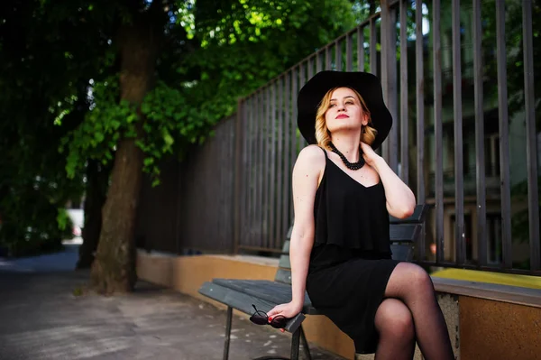 Mujer rubia en vestido negro, collares y sombrero sentado en el banco . — Foto de Stock