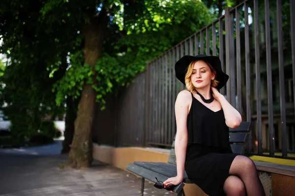 穿着黑色衣服的金发碧眼的女人, 坐在长凳上的项链和帽子. — 图库照片