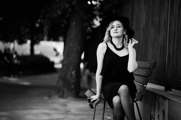 Siyah elbise, kolyeler ve şapkasını bankta oturan sarışın kadın. — Stok fotoğraf