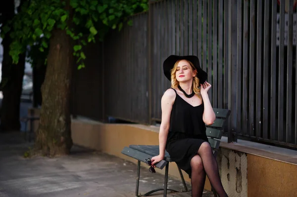 穿着黑色衣服的金发碧眼的女人, 坐在长凳上的项链和帽子. — 图库照片