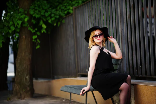 Blonde woman on black dress, necklaces and hat sitting on bench. — Stock Photo, Image