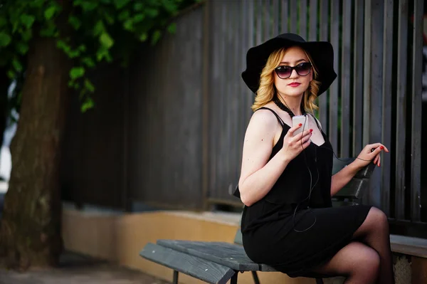 Blonde woman on black dress, sunglasses, necklaces and hat sitti — Stock Photo, Image