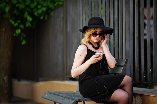 黑色礼服, 太阳镜, 项链和帽子坐姿的金发碧眼的女人 — 图库照片