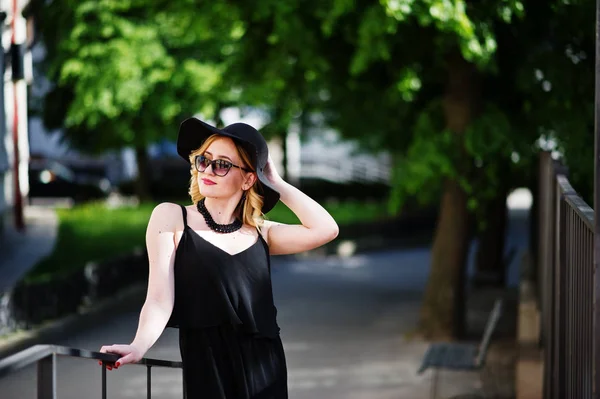 Blond kvinna på svart klänning, solglasögon, halsband och hatt ställde — Stockfoto
