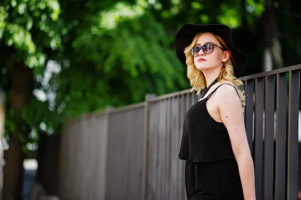 Donna bionda su abito nero, occhiali da sole, collane e cappello in posa — Foto Stock