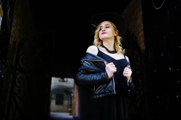 Mujer rubia en vestido negro, chaqueta de cuero, collares y sombrero a — Foto de Stock