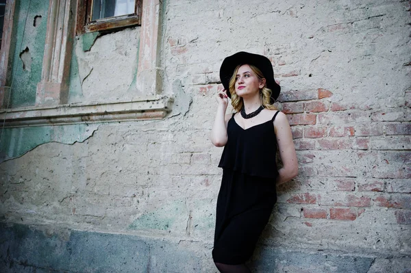 Femme blonde sur robe noire, colliers et chapeau contre vieux mur — Photo