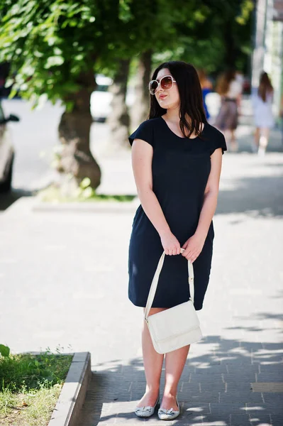 Brunett tjej på svart klänning på solglasögon med handväska till hands — Stockfoto
