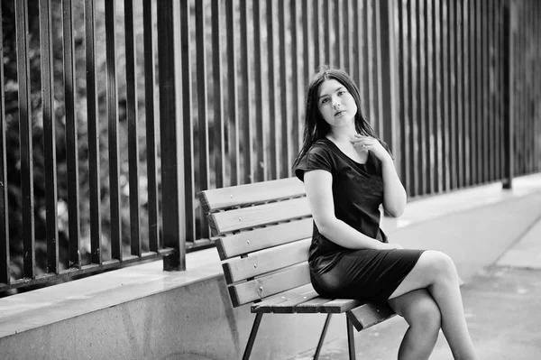 Brünettes Mädchen im schwarzen Kleid sitzt auf Bank und posiert bei stre — Stockfoto