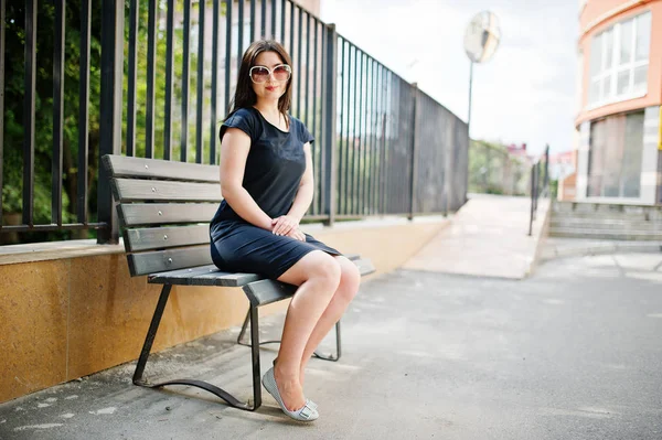 Brunette fille à robe noire sur les lunettes de soleil assis sur le banc et — Photo