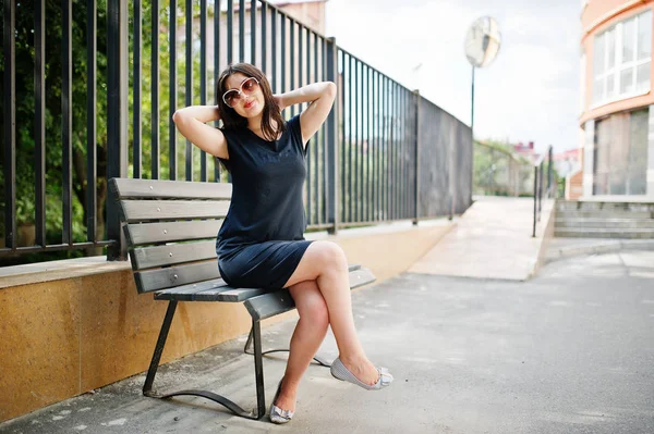 Brünettes Mädchen im schwarzen Kleid mit Sonnenbrille auf Bank sitzend und — Stockfoto