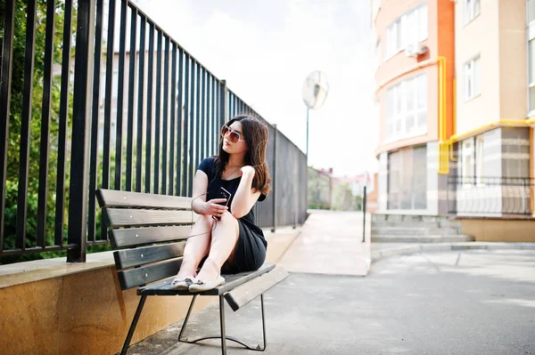 Brunett tjej på svart klänning, solglasögon sitter på bänken, liste — Stockfoto
