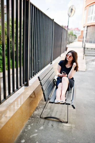 Menina morena no vestido preto, óculos de sol sentados no banco, liste — Fotografia de Stock
