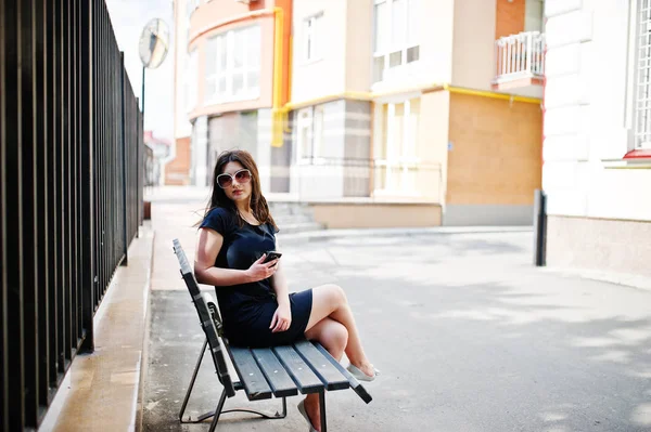 Brunett tjej på svart klänning, solglasögon sitter på bänken, liste — Stockfoto