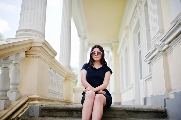 Brunett tjej på svart klänning, solglasögon sitter på trappan till ol — Stockfoto
