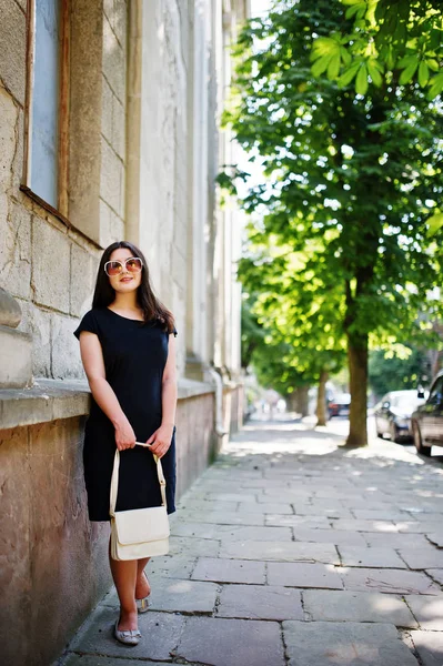 Brünettes Mädchen im schwarzen Kleid mit Sonnenbrille und Handtasche in der Hand — Stockfoto