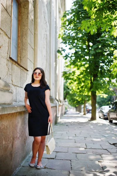 Brünettes Mädchen im schwarzen Kleid mit Sonnenbrille und Handtasche in der Hand — Stockfoto