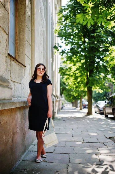 Brünettes Mädchen im schwarzen Kleid mit Sonnenbrille und Handtasche in der Hand — Stockfoto